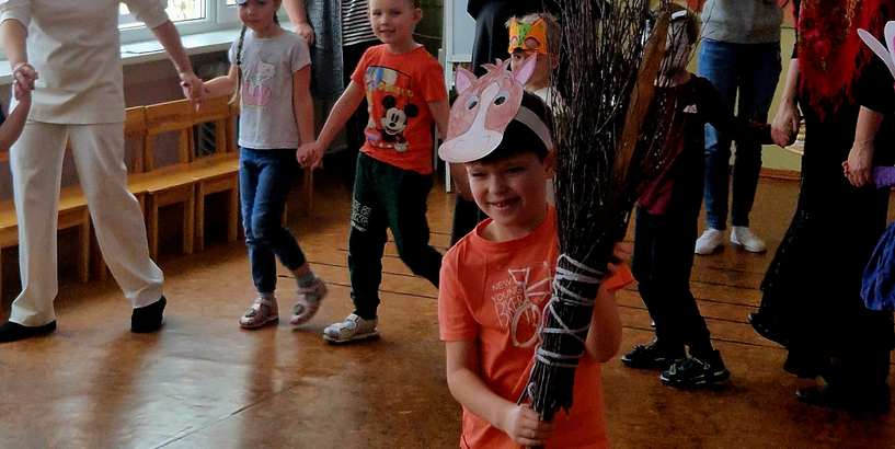 Bērni, sadevušies rokās, iet  Meteņdienas rotaļā, centrā dalībnieks dejo ar zaru slotu