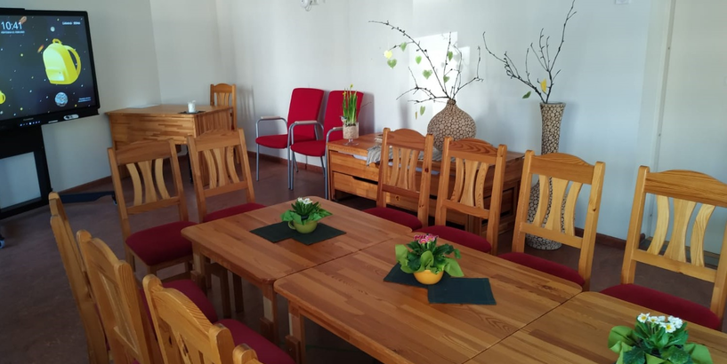 Rīgas 275. pirmsskolas izglītības iestādes “Austriņa” pasākuma telpa ar koka galdu, krēsliem un ekrānu