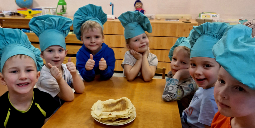 Bērnudārza grupiņas bērni apsēdušies ap galdu pavāru cepurēs pēc pīrādziņu cepšanas