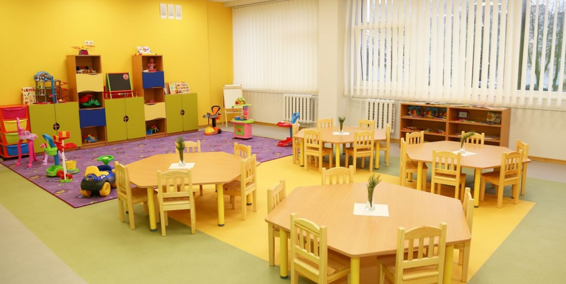 Bērnudārza grupiņas telpa ar galdiem, krēsliem un rotaļu stūrīti