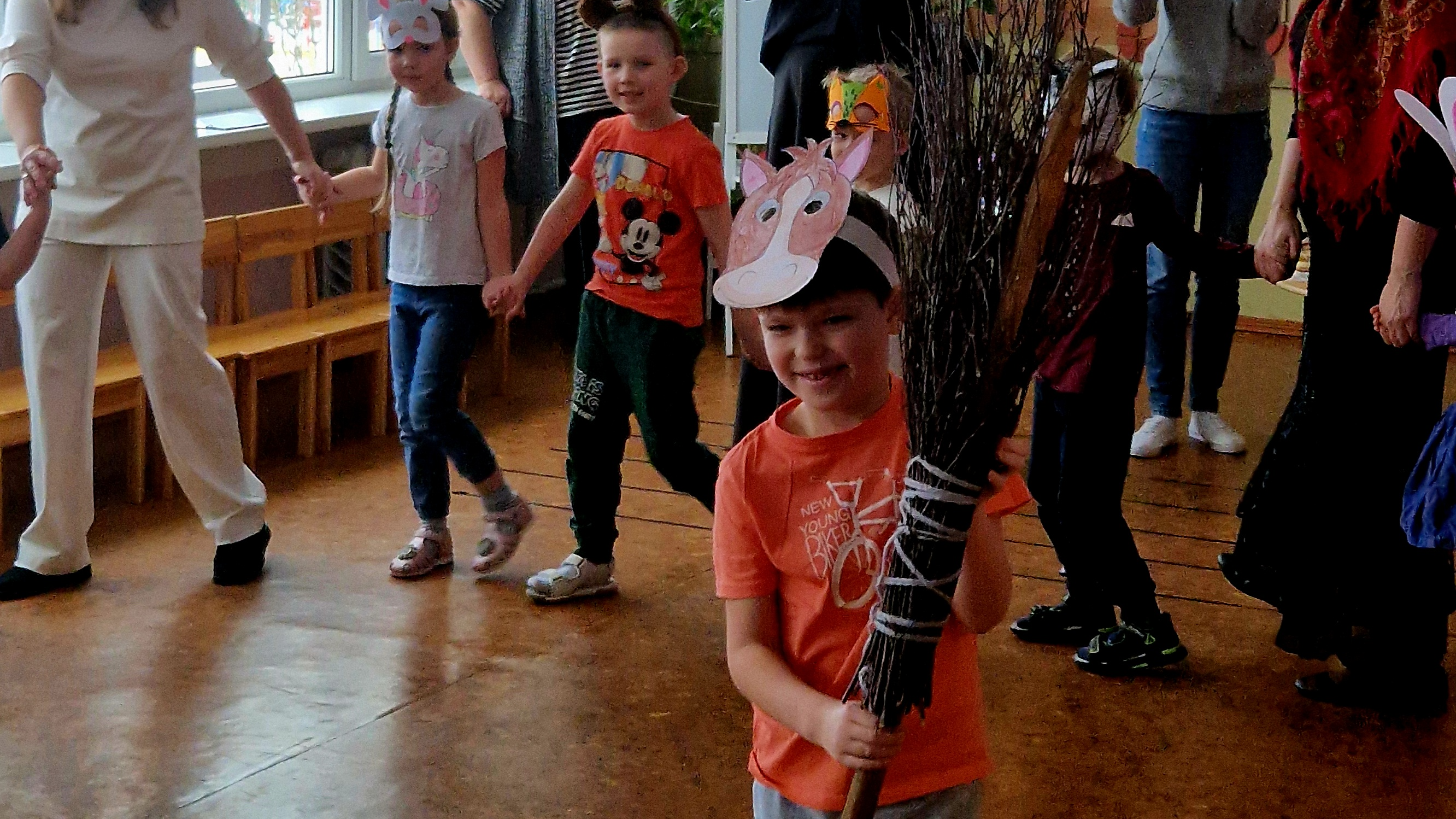 Bērni, sadevušies rokās, iet  Meteņdienas rotaļā, centrā dalībnieks dejo ar zaru slotu